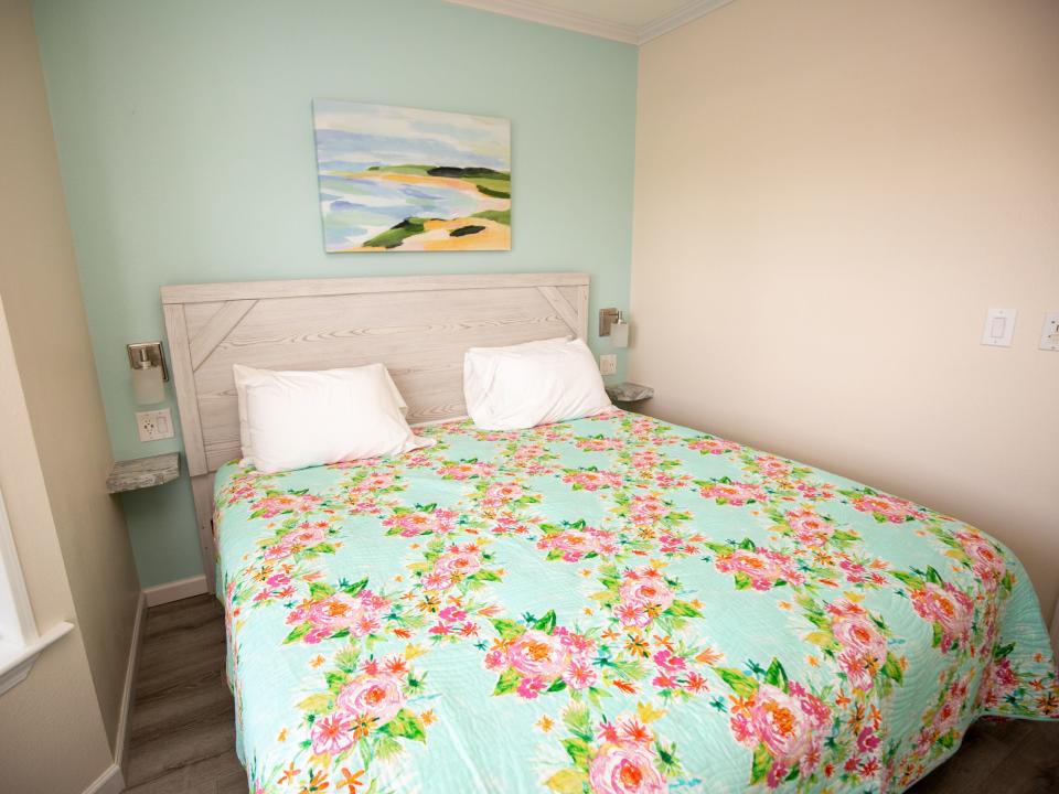 A bed with a flower pattern on the blanket. There's a picture on the wall.