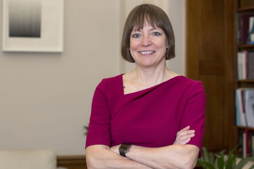 Professor Shearer West is a white woman of slim build with short brown hair and blue eyes. In this picture, she is standing smiling at the camera with her arms crossed. She is wearing fuchsia dress