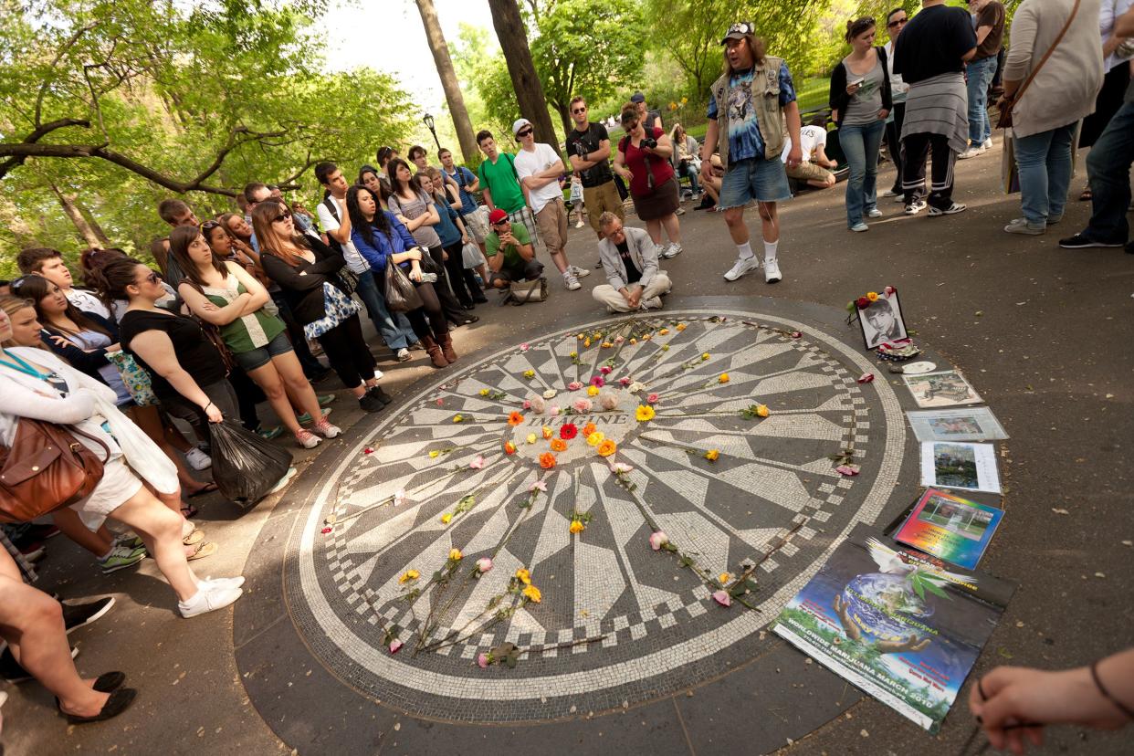 John Lennon's Strawberry Fields Monument in Central Park, New York City, where his ashes were scattered, several people surround the 'Imagine' circle with brightly colored flowers on it and pictures on the side with trees in the background in summer
