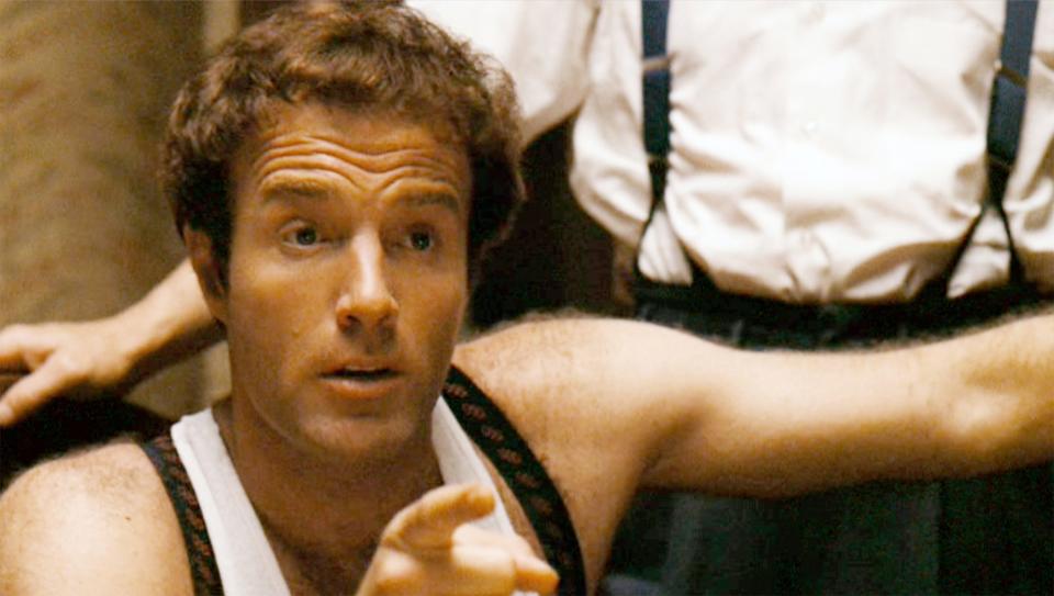 Caan in character pointing and wearing a a white tank top and suspenders.