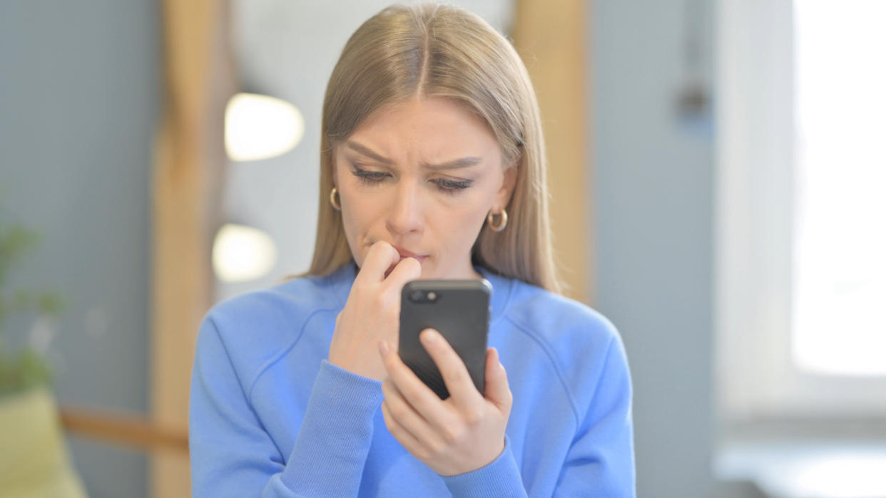 A young woman looks worriedly at her smartphone while biting her nails