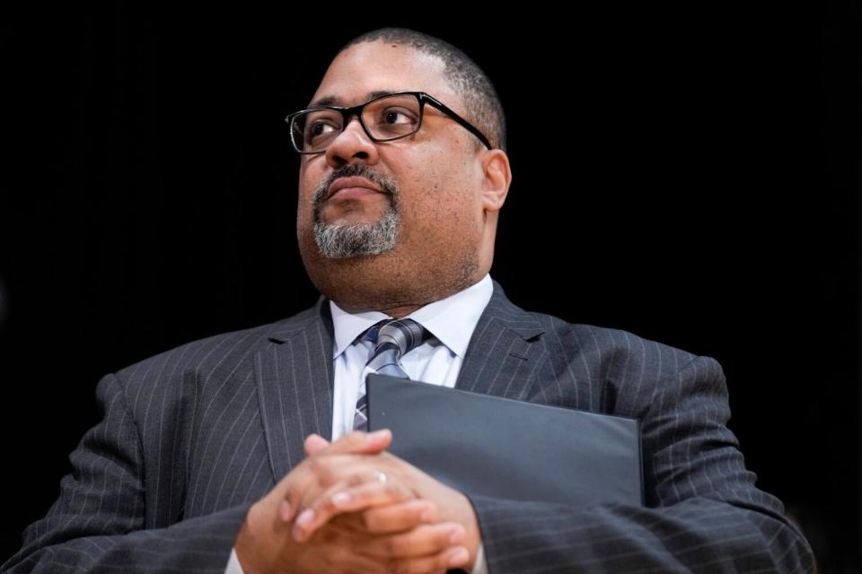 A lower-angle head-and-shoulders horizontal frame of Alvin Bragg wearing a striped dark suit, tie and black-frame glasses