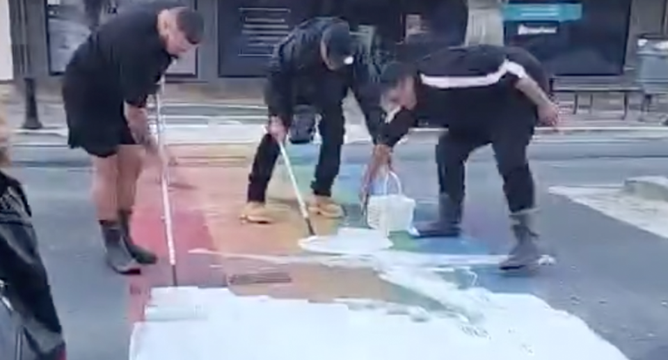 Three men work together to cover white paint onto the rainbow crossing with paint rollers. 