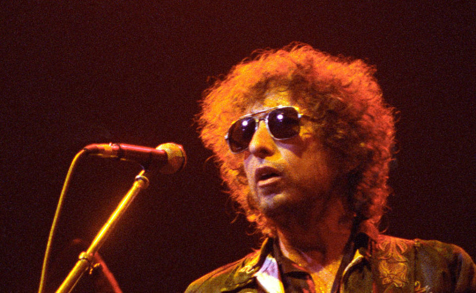 Bob Dylan singing into a microphone while wearing sunglasses and a casual outfit