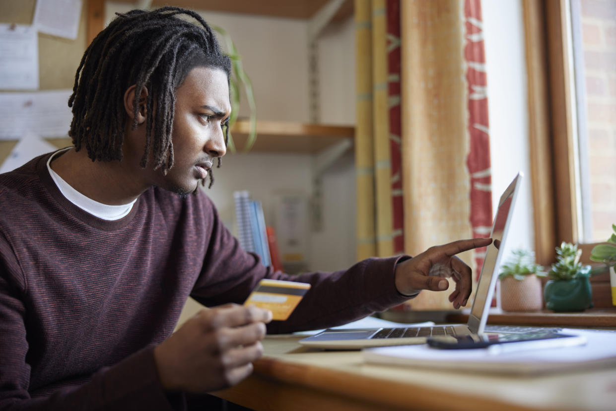 Male University Or College Student With Credit Card Looking At Laptop Worried About Debt At Desk In Room