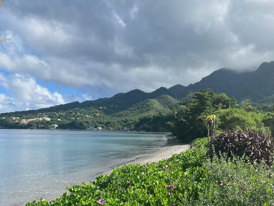 Greenery and sand next to beautiful blue waters. There are hazy, greenery-covered mountains in the background.