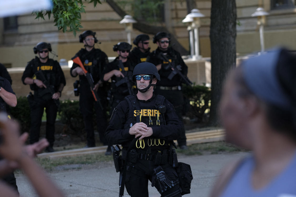 Police in riot gear stand by as demonstrators gather.