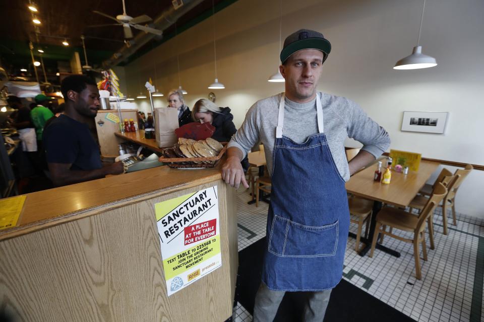 In this Wednesday, Jan. 18, 2017 photo, Ben Hall, owner of the Russell Street Deli, poses with a sanctuary restaurant sign in Detroit. Dozens of restaurants are seeking “sanctuary” status, a designation owners hope will help protect employees in an immigrant-heavy industry and tone down fiery rhetoric sparked by the presidential campaign. (AP Photo/Paul Sancya)