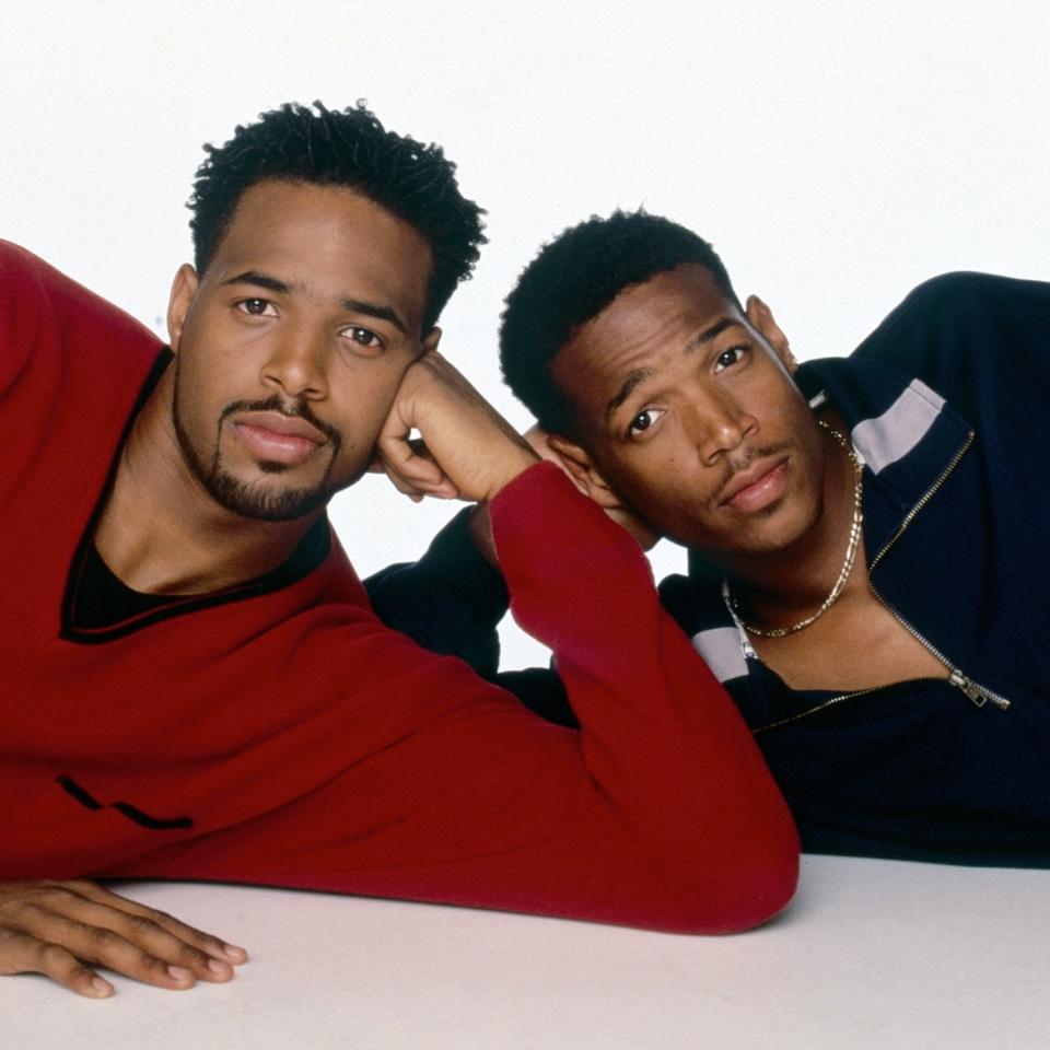 Photo of Shawn Wayans in a red sweater and Marlon Wayans in a black jacket with his hand resting on his face, both reclining and gazing at the camera