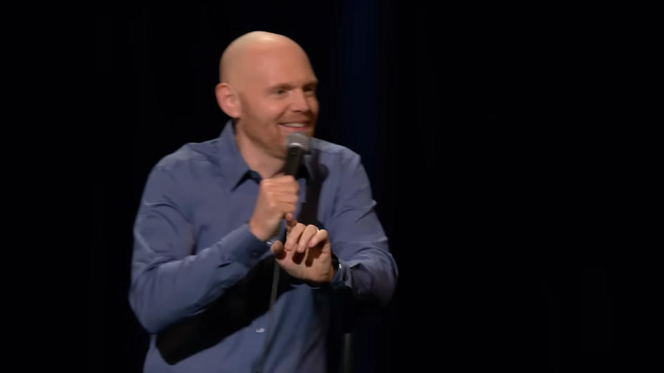 Bill Burr smiling in a blue shirt while on stage.