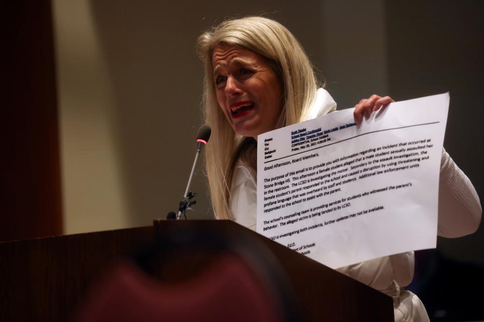 Heather Fruzzetti speaks into a microphone at a podium holding a printed piece of paper.