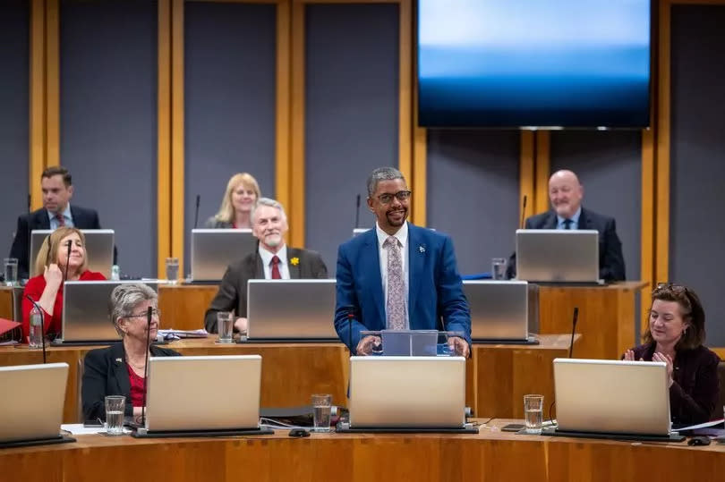 Vaughan Gething smiling in the Senedd, standing up while being applauded by Labour MSs previously