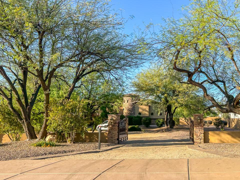 An open gate in a driveway leads to a castle-like house shaded by several trees