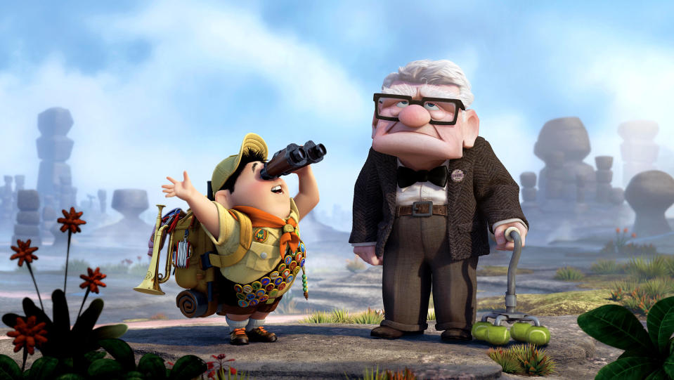 Russell, a young boy scout with binoculars and a backpack, stands next to Carl Fredricksen, an elderly man with a cane, in a rocky landscape from "Up."