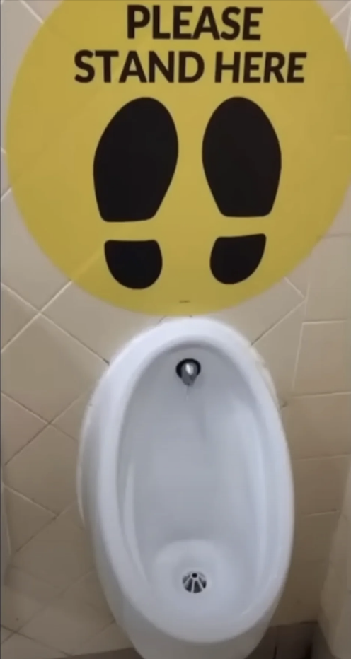 Sign above urinal reads "PLEASE STAND HERE" with footprints indicating where to stand