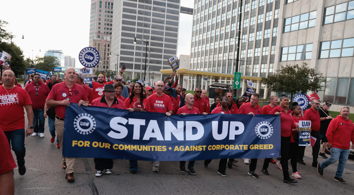 United Auto Workers president Shawn Fain and UAW members march in the street. They hold a banner reading: Stand up for our communities, against corporate greed.