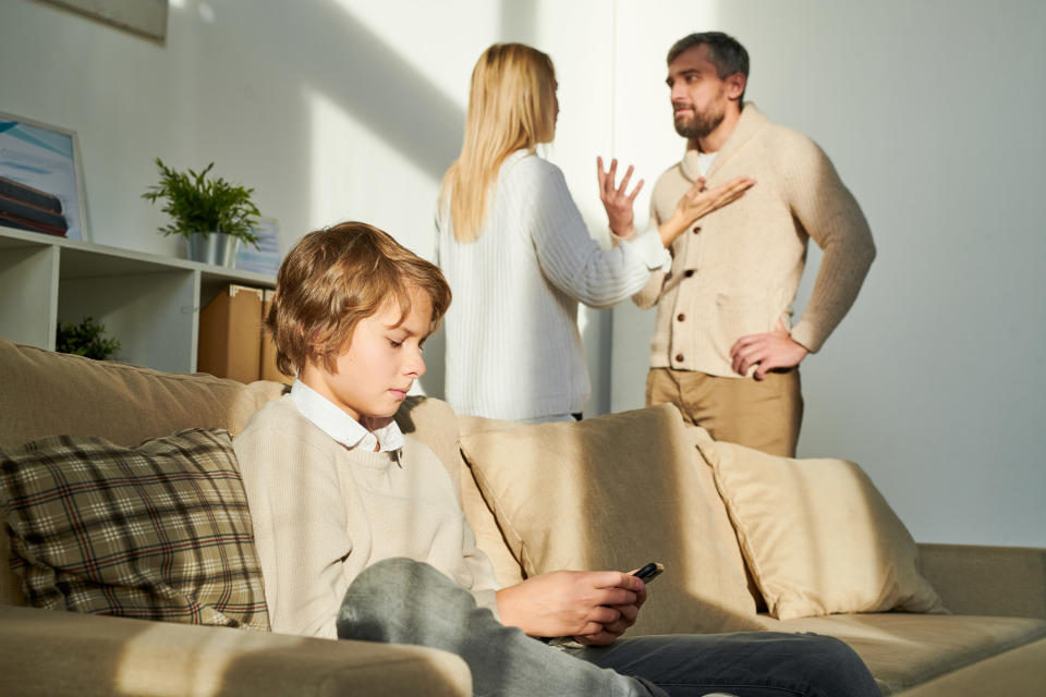 Serious concentrated teenage boy being closed off sitting on comfortable sofa and using smartphone while parents having quarrel in background