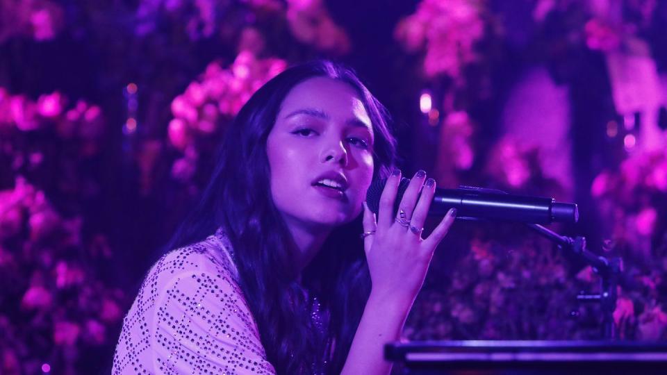 olivia rodrigo sits at a piano and sings into a microphone, she wears several rings and a bejeweled shirt