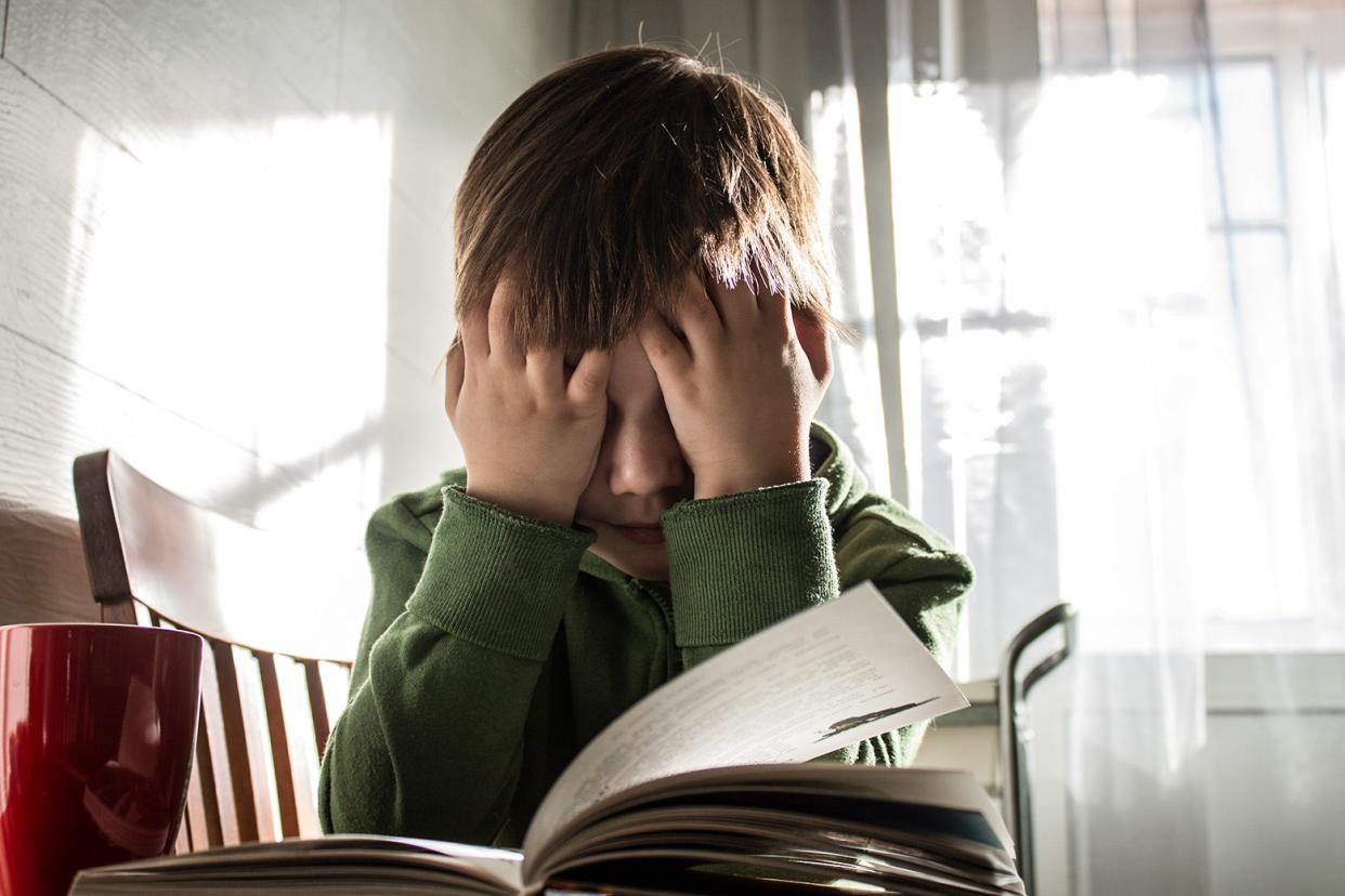 A frustrated child sits at a table, their head in their hands, with a school book open.
