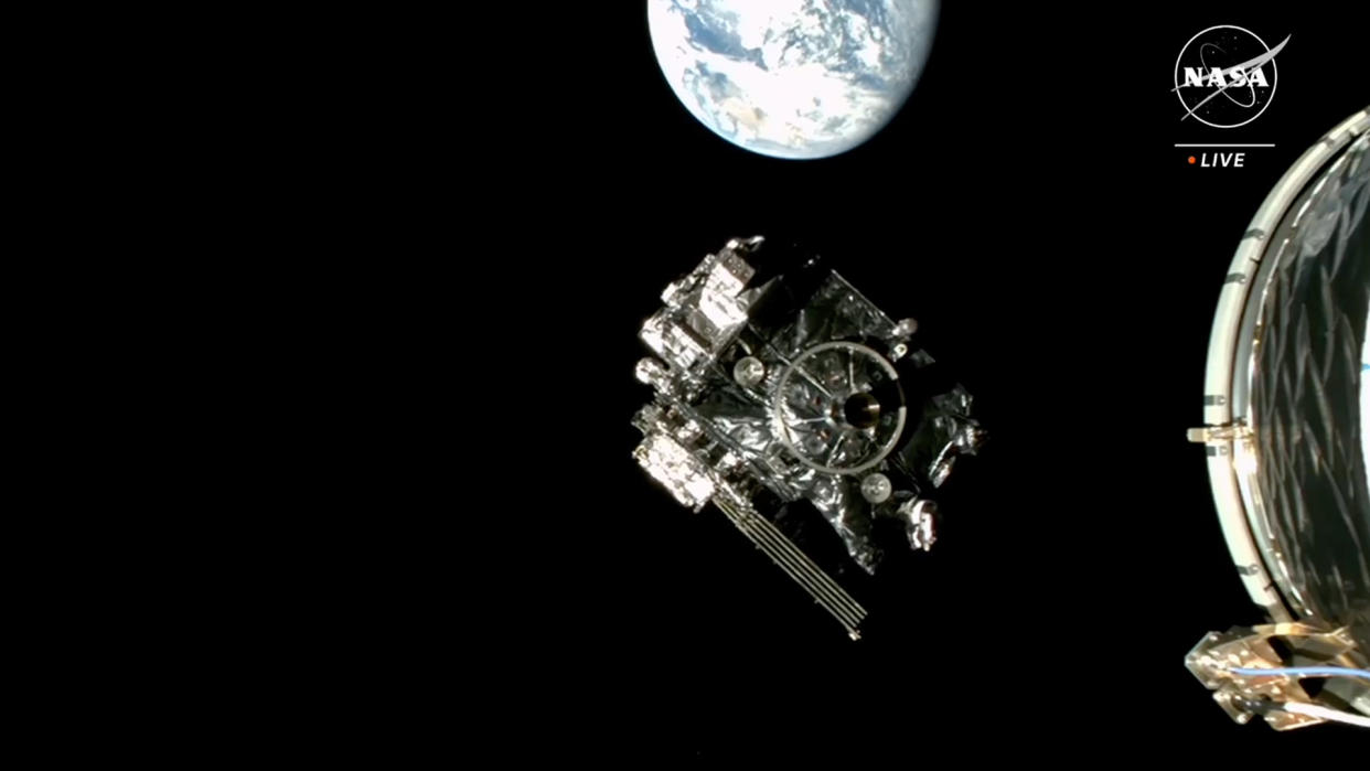  A cube-shaped spacecraft can be seen against the blackness of space. a bright blue-and-white earth can be seen in the background. 