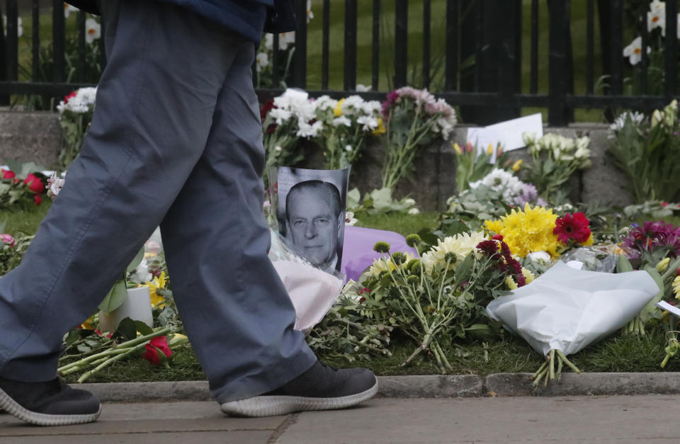 A man walks past a photo of Britain's Prince Philip among flowers left by the public outside the gates of Windsor Castle, a day after the death of Britain's Prince Philip, in Windsor, England, Saturday, April 10, 2021. Britain's Prince Philip, the irascible and tough-minded husband of Queen Elizabeth II who spent more than seven decades supporting his wife in a role that mostly defined his life, died on Friday. (AP Photo/Frank Augstein)