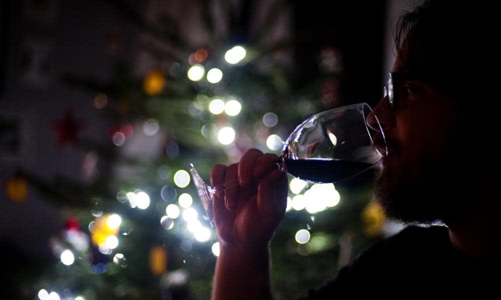 A man drinks a glass of wine by a Christmas tree.