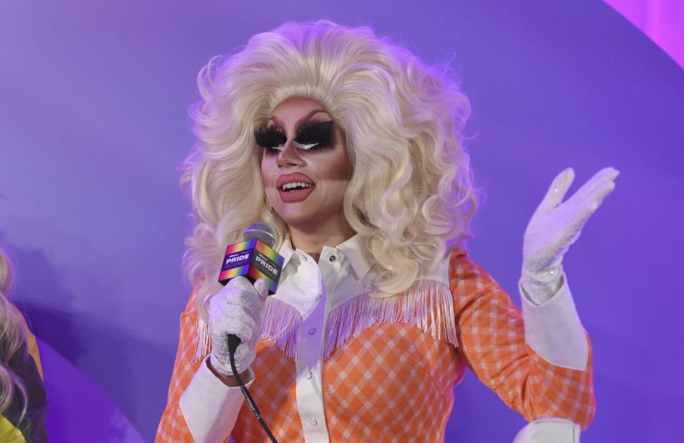 A person wearing a large blond wig and heavy makeup in an orange dress and white gloves