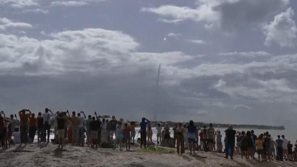 a large white rocket rises through a cloudy sky above a huge plume of fire and smoke