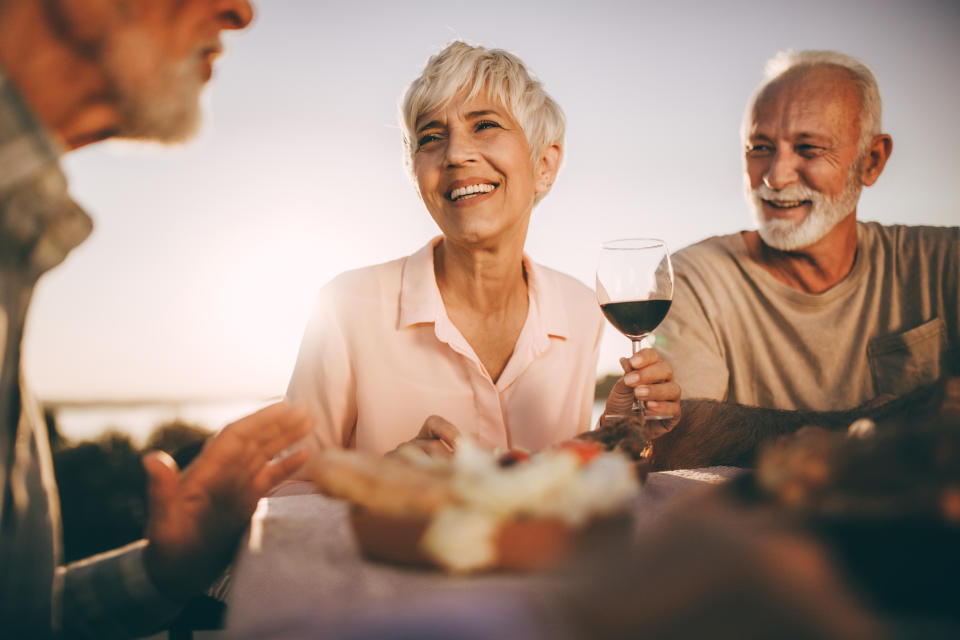 Three older adults sit together, smiling and enjoying a meal outdoors at sunset. One woman holds a glass of wine while the others engage in conversation