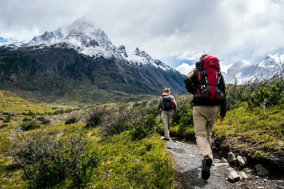 A pair of hikers are moving towards the mountains, they carry packs and are dressed for cooler weather in pants and jackets.