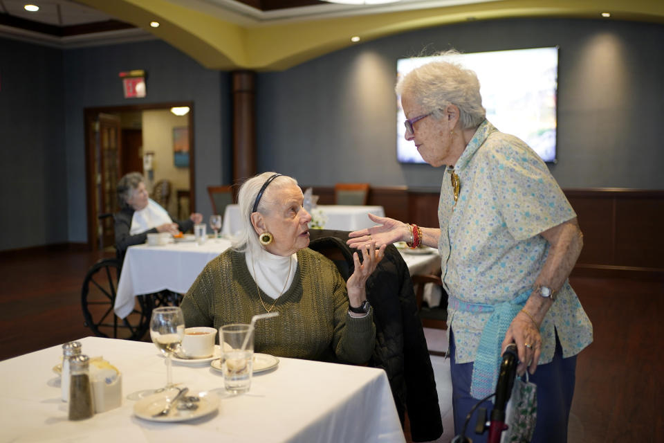 FILE - Ita Aber, right, stops by the table of her old friend Rita Shliselberg after she finished dinner at RiverWalk, an independent senior housing facility, in New York, Thursday, April 1, 2021. Since the start of the pandemic, residents had been dining in their rooms. Only recently have they began to use the dining hall again. A focus on the elderly at the start of the nation's vaccination campaign helped protect nursing homes that were ravaged at the height of the U.S. coronavirus outbreak, but they are far from in the clear. New outbreaks, often traced to infected staff members, are still occurring in long-term care centers across the country, causing continued havoc for visitation policies. (AP Photo/Seth Wenig, File)