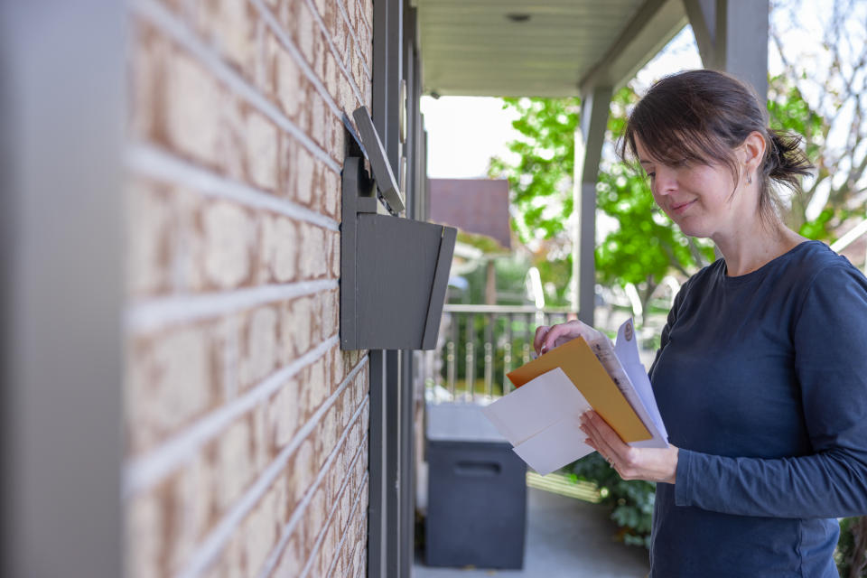 ผู้หญิงคนหนึ่งกำลังรวบรวมไปรษณีย์ที่บ้านในกล่องจดหมายของเธอในออสเตรเลีย เธอกำลังยิ้มและรับจดหมายของเธอ เธอกำลังดูจดหมายที่เธอได้รับ