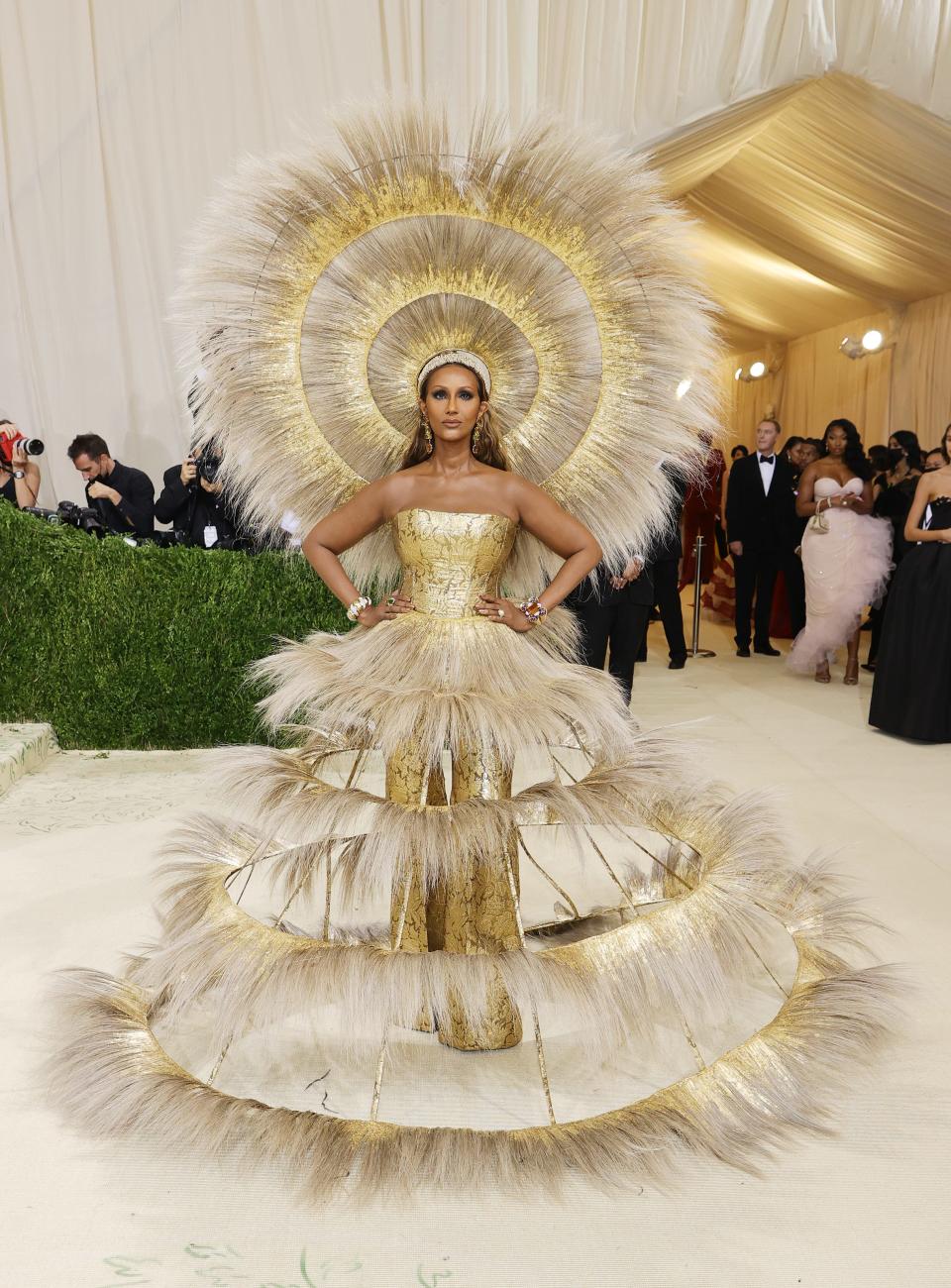 Iman attends The 2021 Met Gala wearing a voluminous gold feathered dress with a large feathered headpiece.