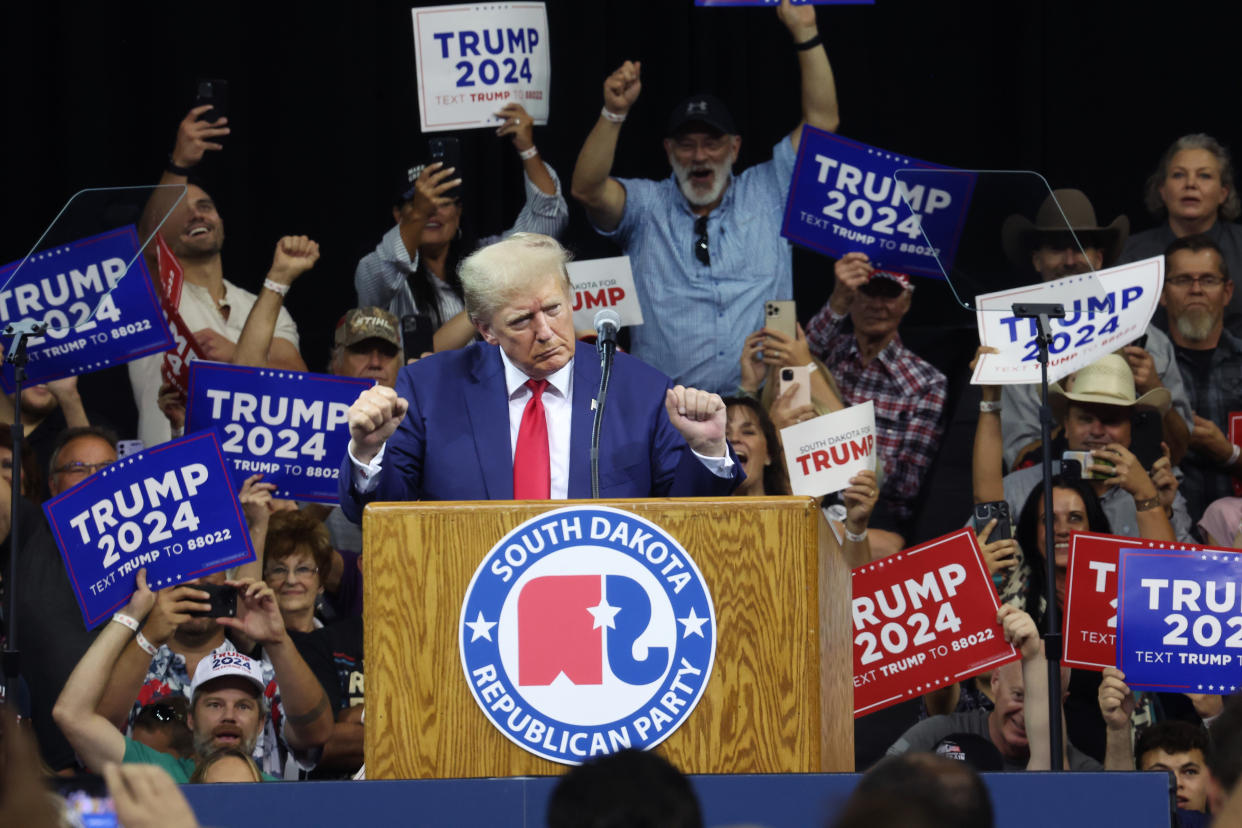 Donald Trump stands at a wooden podium emblazoned with a plaque that reads: South Dakota Republican Party while surrounded by a few dozen people holding signs that read: Trump 2024.