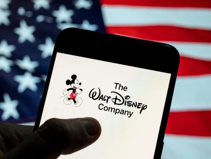 American flag in the background of someone holding a phone that says The Walt Disney Company on the screen.