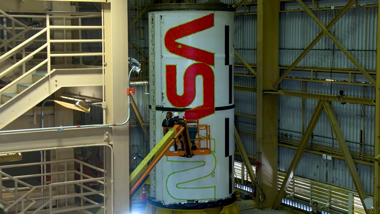  A rocket standing up with a nasa logo half-painted. people on a crane wield spray paint. the rocket is in a warehouse. the door is visible at right and a large staircase visible at left. 