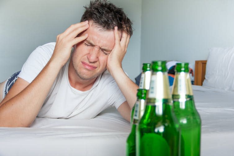 Man lies in bed, clutching his head, with beer bottles in view.