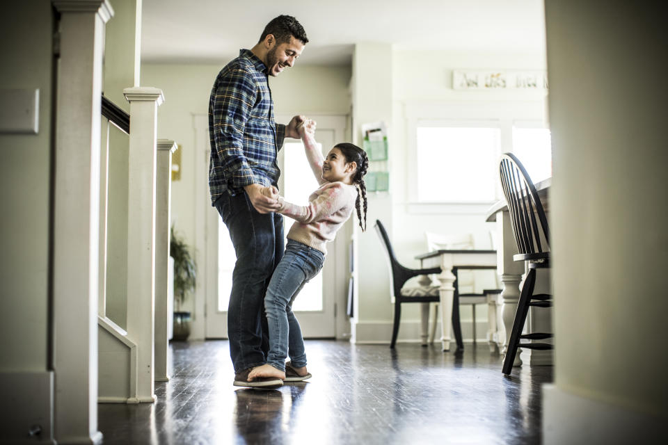 A man dances with his daughter, who is playfully standing on his feet, at home