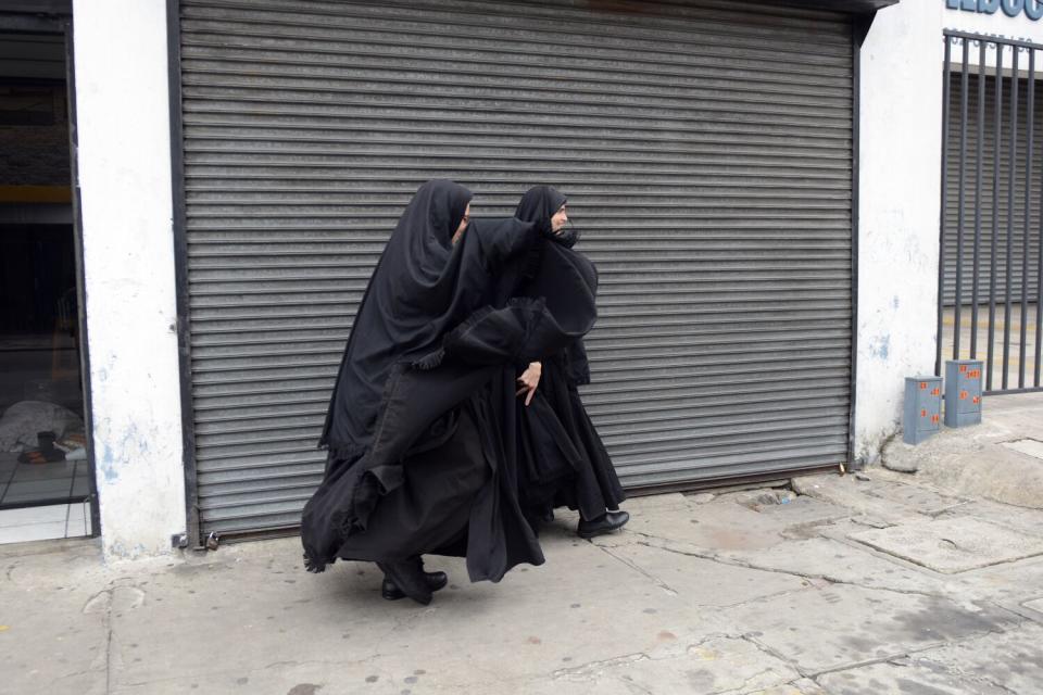 Two women cloaked in black walk on a street past a metal door