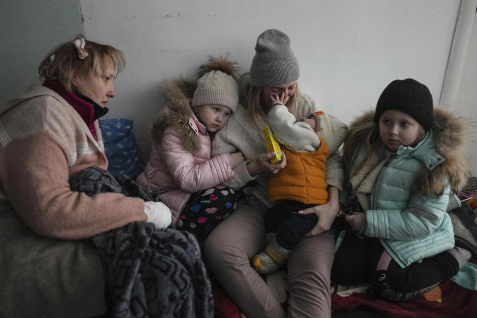 Two women and two children sit on the floor against a white wall.