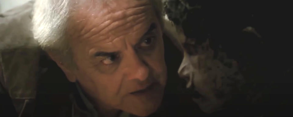 An older man with gray hair, in a tense scene from a movie or TV show, closely faces a person with a distorted face of a boy