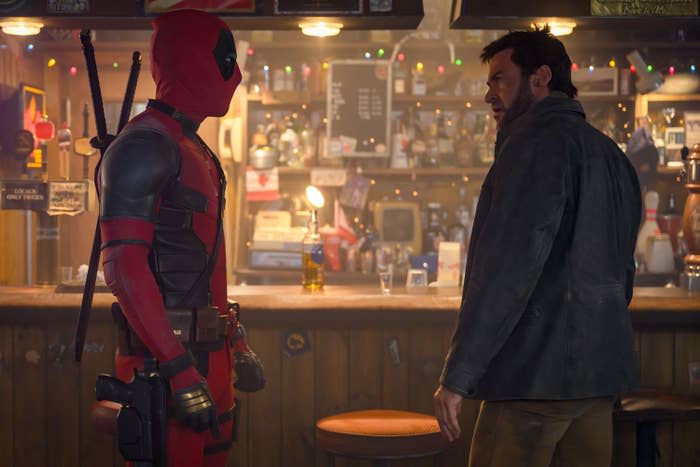 Ryan Reynolds as Deadpool in costume and Hugh Jackman facing each other in a character-filled bar scene with festive lights and bottle-filled shelves