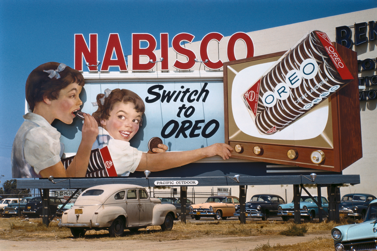 Vintage Nabisco Oreos billboard ad surrounded by parked cars, two girls eating Oreos while watching TV with Oreos on the TV screen, brown dry grass and a clear blue sky, 1950s