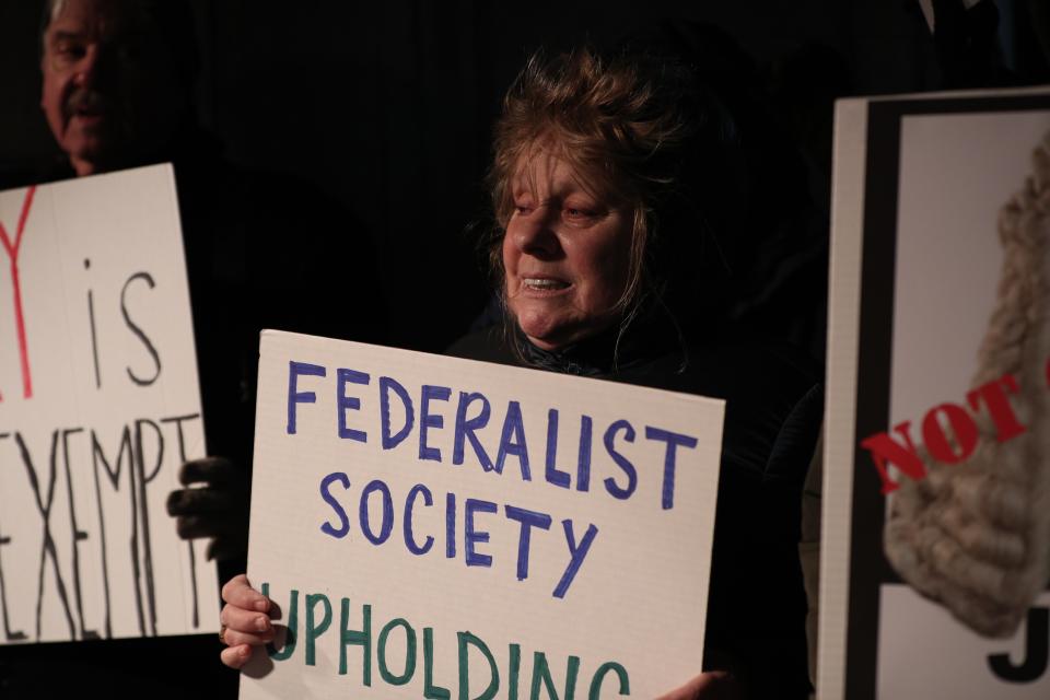 A middle-aged woman holds a placard saying: Federalist Society Upholding...