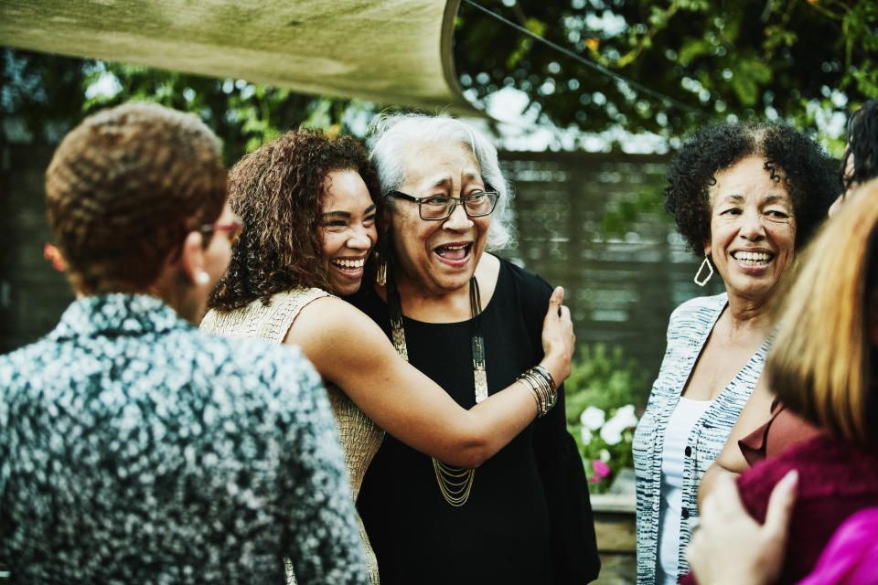A group of women, including an elderly woman being hugged, are outdoors under a canopy, smiling and engaging in a joyful conversation
