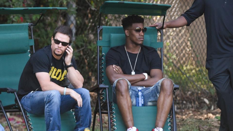 mark wahlberg and jimmy butler, both wearing black t shirts, jeans, and sunglasses, sit on green lawn chairs outside on the grass