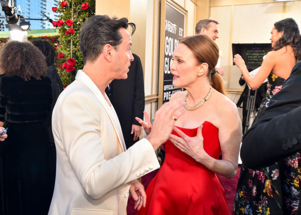 A man in a white suit and a woman in a red gown are in conversation on the red carpet at an event. Other attendees are visible in the background