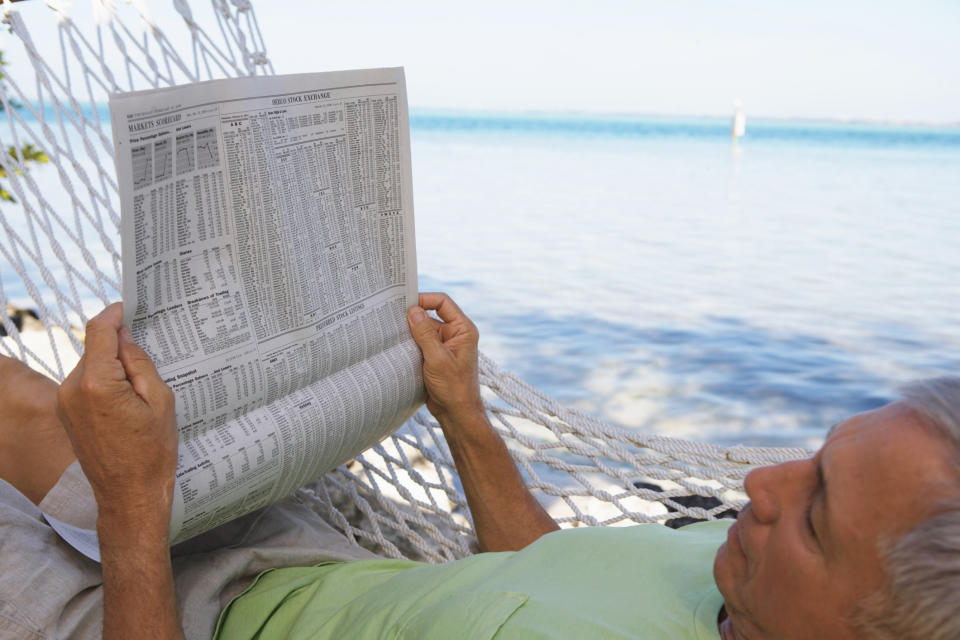 A man resting in a hammock near the ocean reading a newspaper.