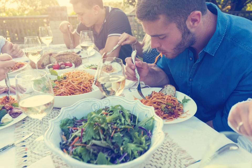 Group of friends having a meal outdoors. All are hungry and eating. Candid shot.  There are bowls of food on the table including salad and spaghetti Bolognese. There are also glasses of wine.  Multi ethnic group.