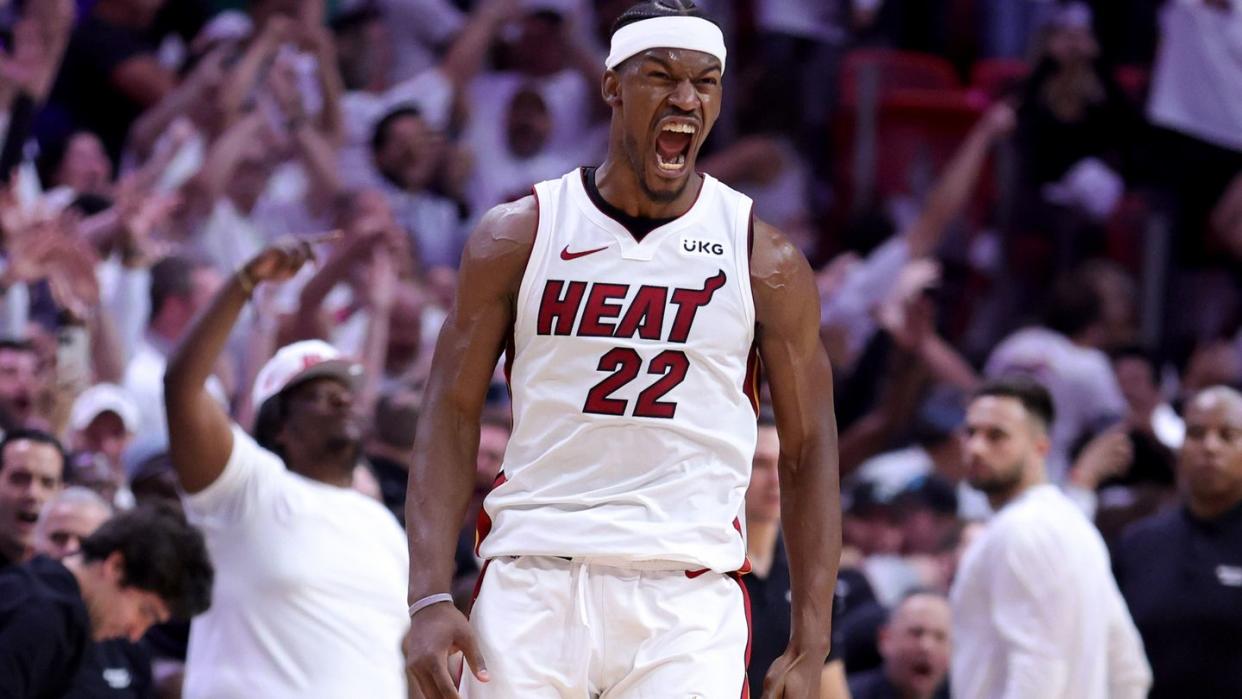 jimmy butler wearing a white miami heat jersey, cheering in excitement, with a crowd in the basketball court stands behind him